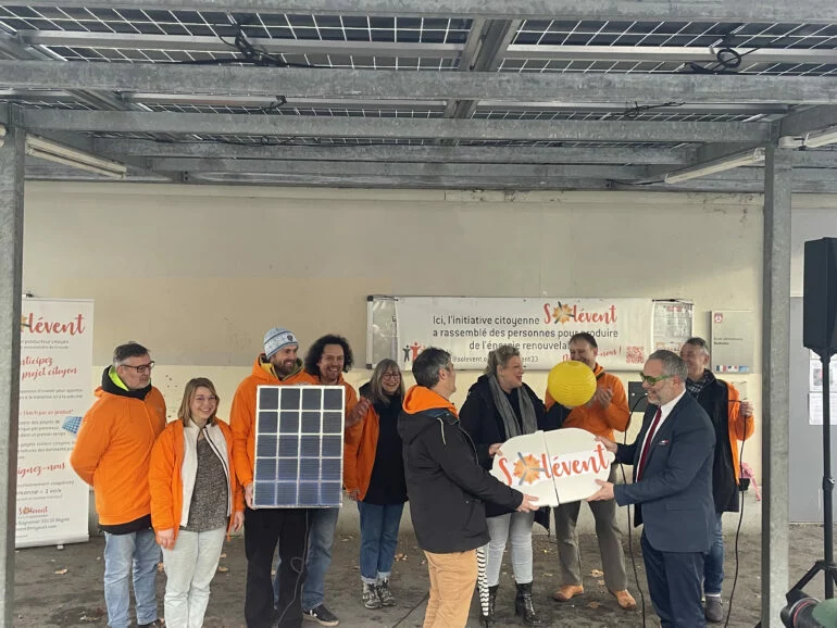 Bénévoles de Solévent en gilets orange tenant un panneau solaire et le logo de la coopérative sous une ombrière photovoltaïque