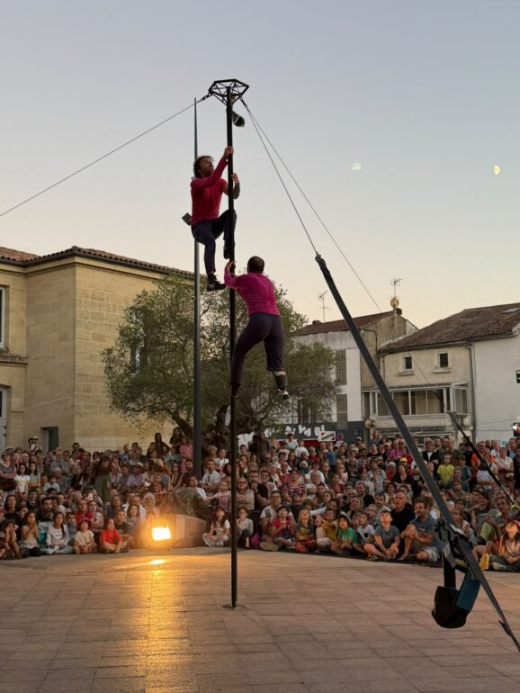 Deux artistes de la compagnie BaNCALE en haut d'un mât, place des Carmes à Langon, devant le public assis.